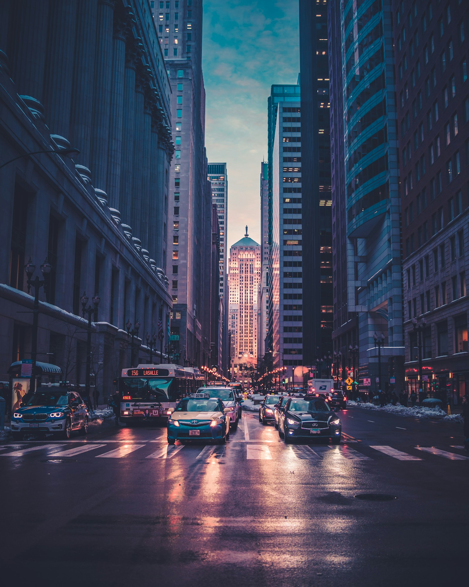Avenida entre rascacielos al atardecer, con el asfalto mojado y luces azules y rosas